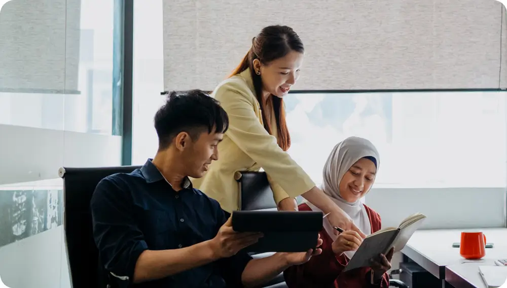 3 people in a meeting referring to data from a book and tablet